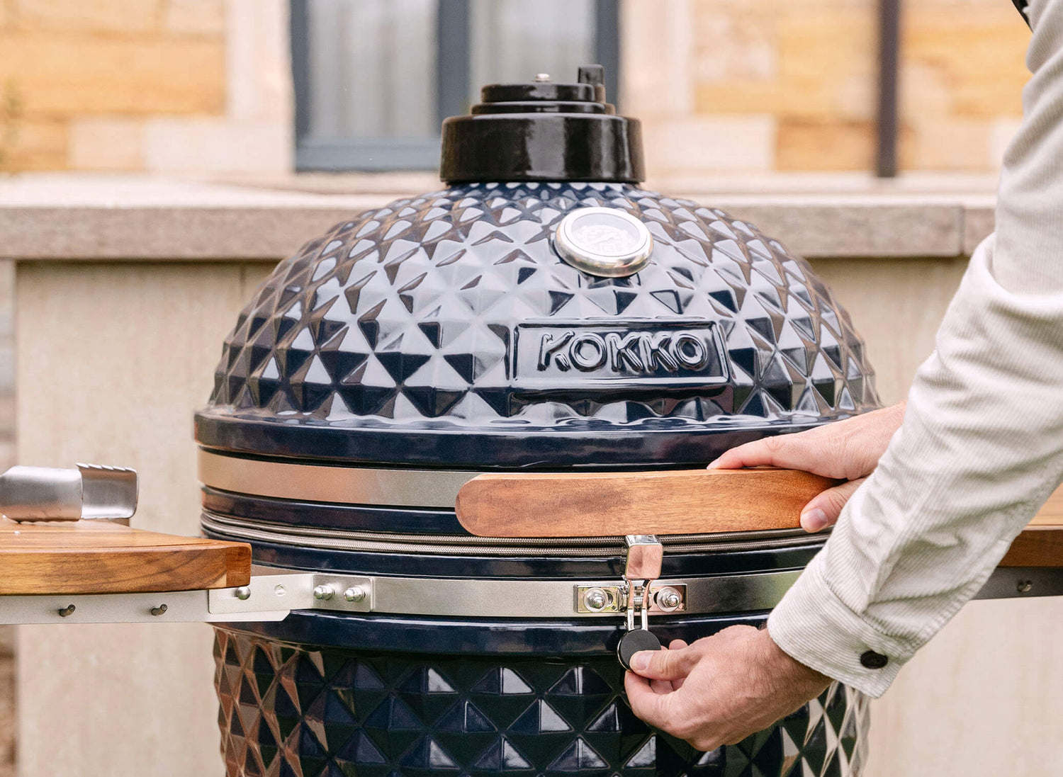 Mano che aziona il fermo di sicurezza di un kamado Kokko nero, con coperchio in ceramica testurizzata, maniglia in legno e termometro integrato, durante l’uso all’aperto.