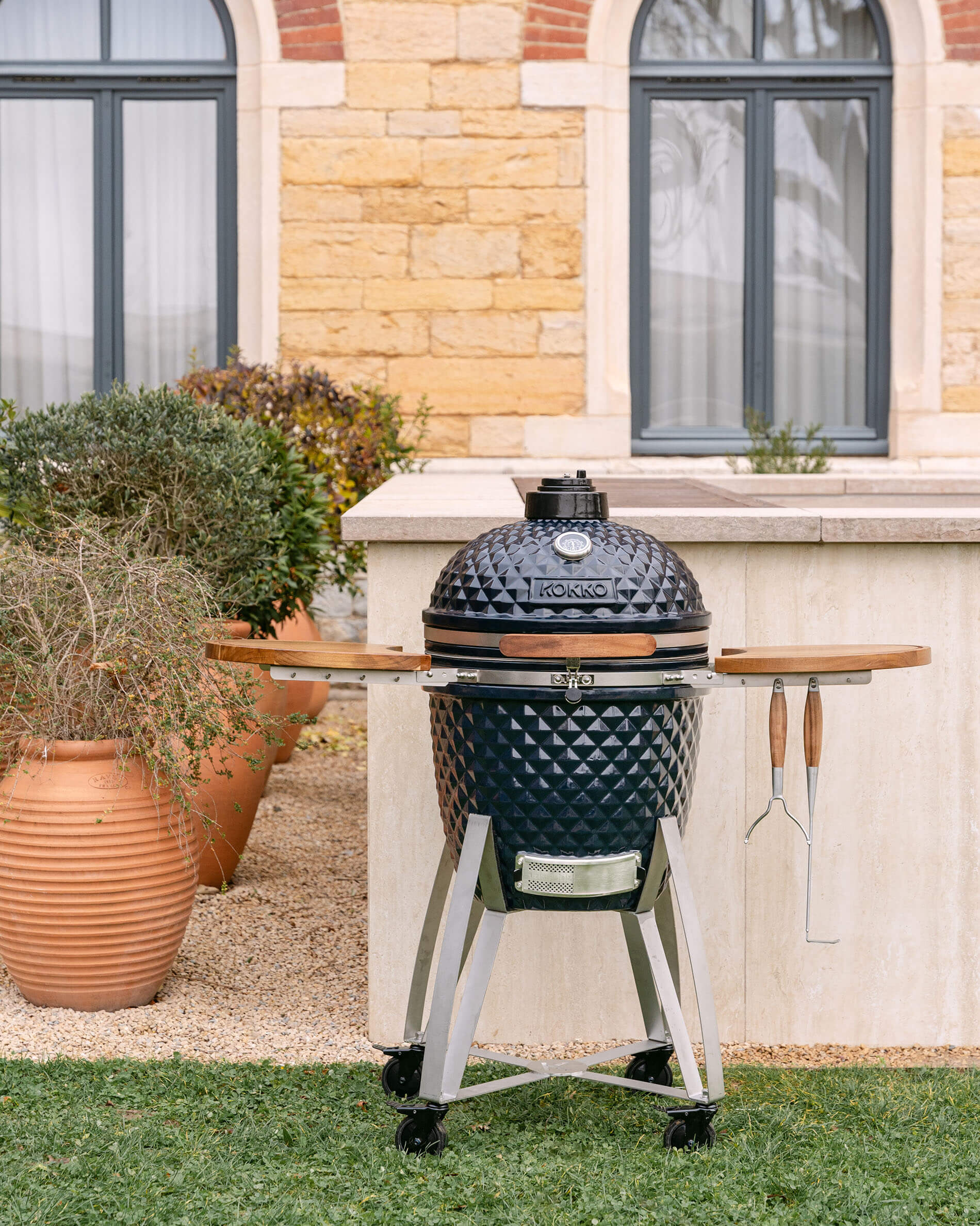 Vista d’insieme del kamado Kokko su carrello mobile con ripiani pieghevoli e utensili appesi, in ambiente esterno.