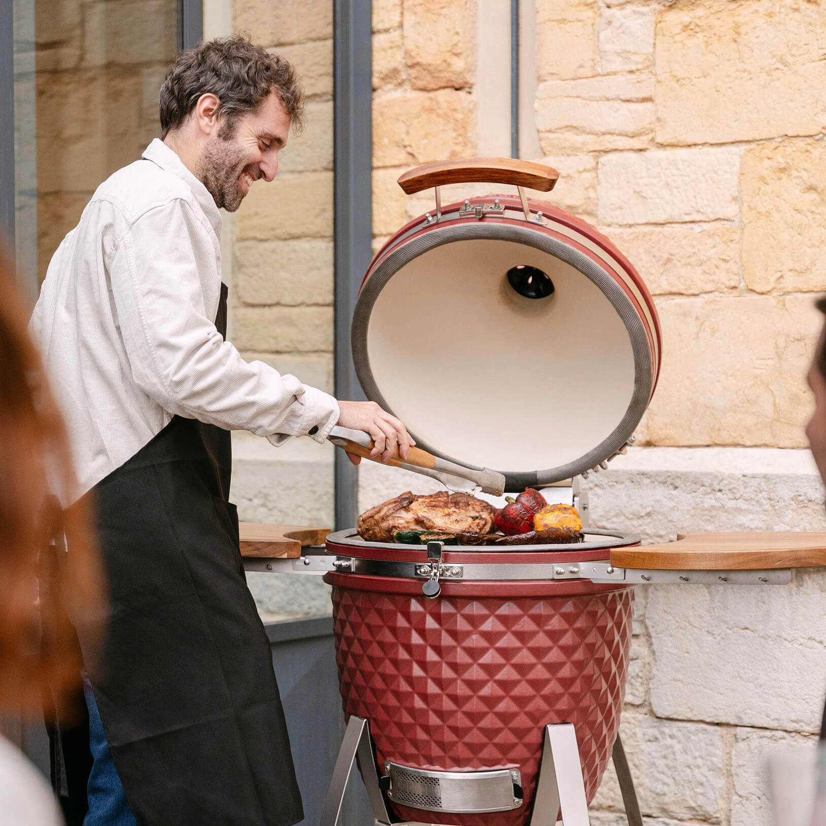 Un utente cucina carne e verdure su un kamado Kokko rosso in ceramica, con il coperchio aperto, durante un momento conviviale all’aperto.
