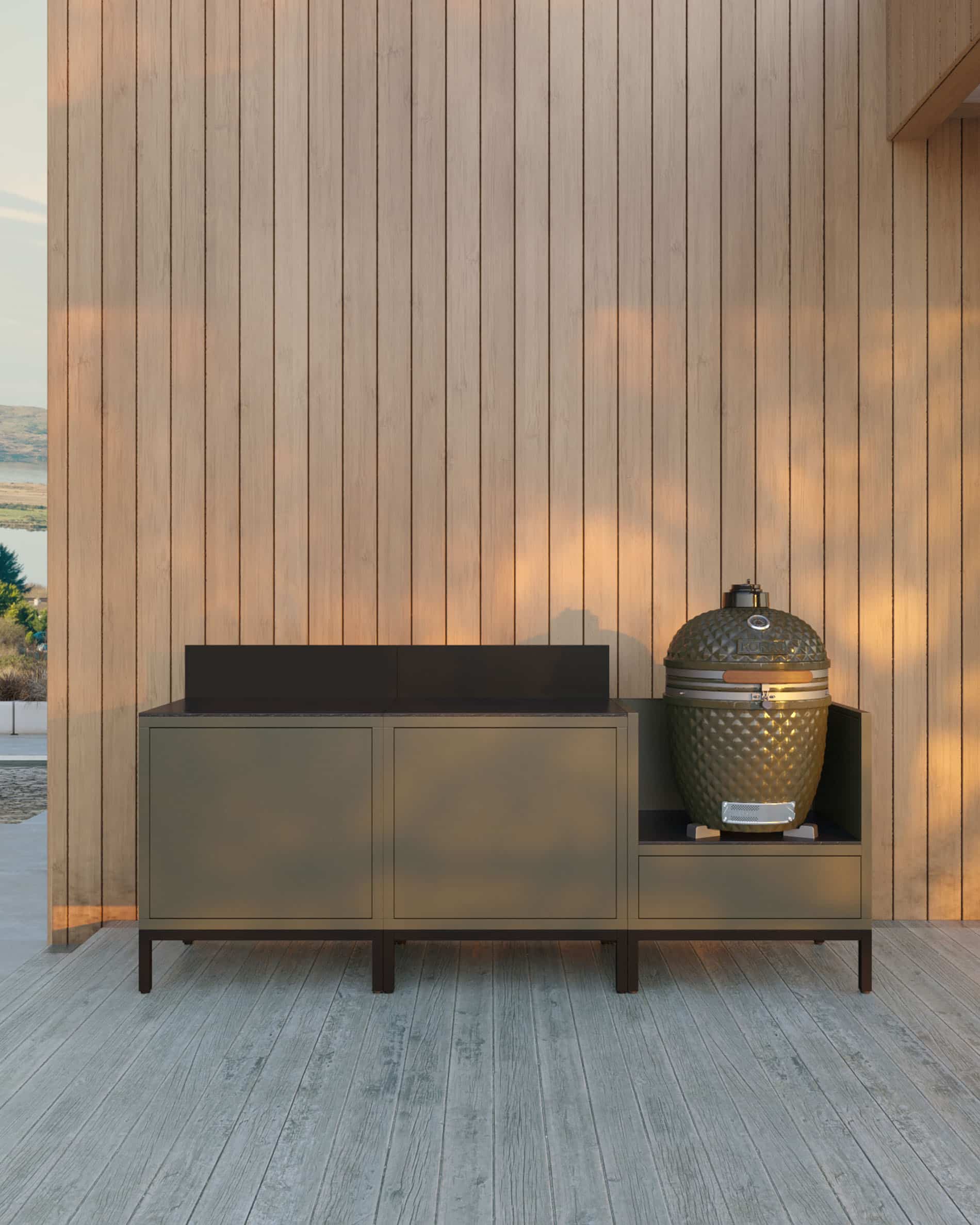 Cucina su pilotis a 3 moduli per kamado, mobile color verde oliva con piano di lavoro scuro e alzatina coordinata, due moduli contenitori chiusi a sinistra e kamado integrato nel modulo laterale destro, struttura su piedini neri, installazione all’esterno contro una parete in legno.