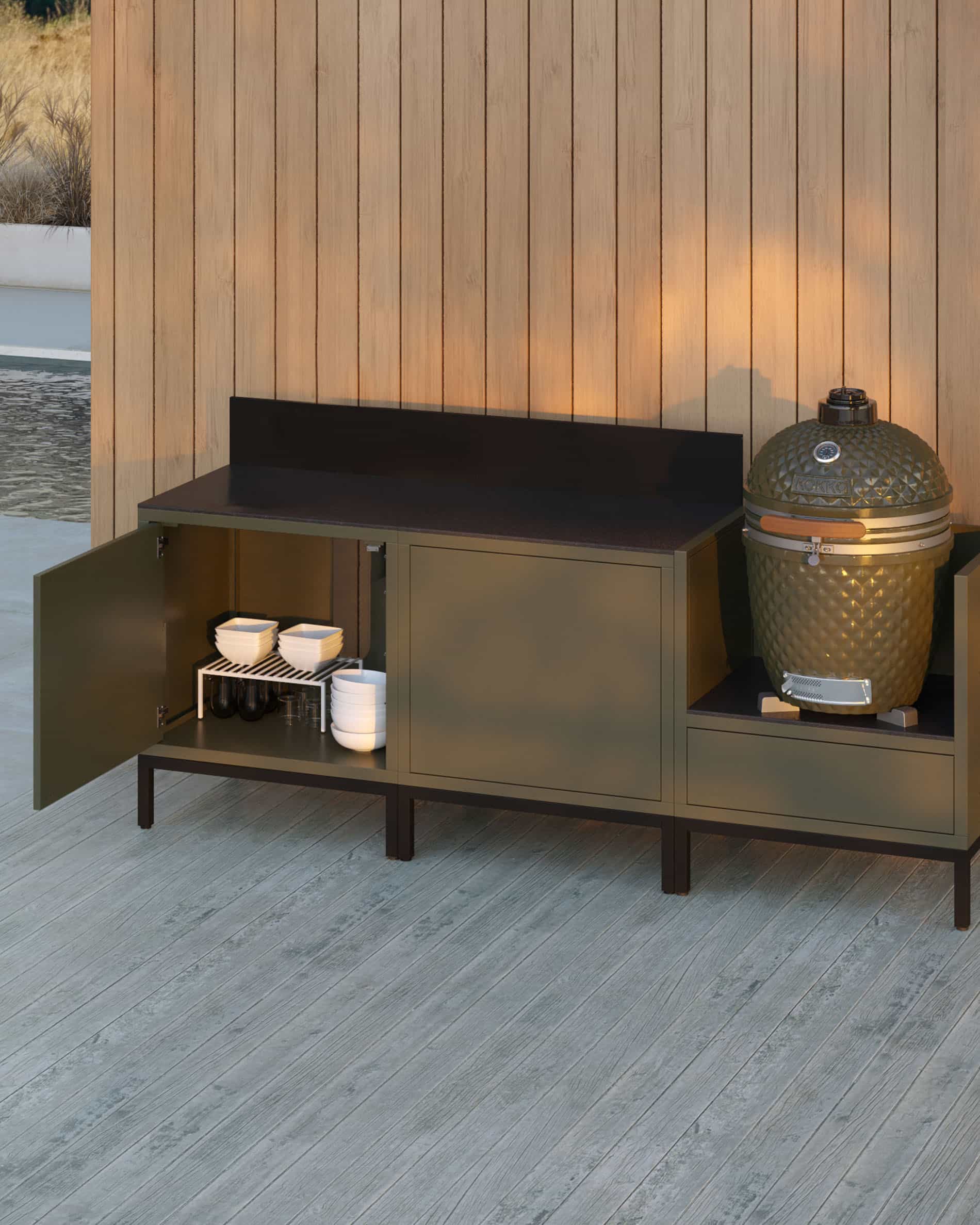 Cucina su pilotis a 3 moduli per kamado, mobile color verde oliva con piano di lavoro scuro e alzatina coordinata, anta del vano contenitore aperta che mostra stoviglie riposte, kamado installato nel modulo laterale destro, struttura su piedini neri.