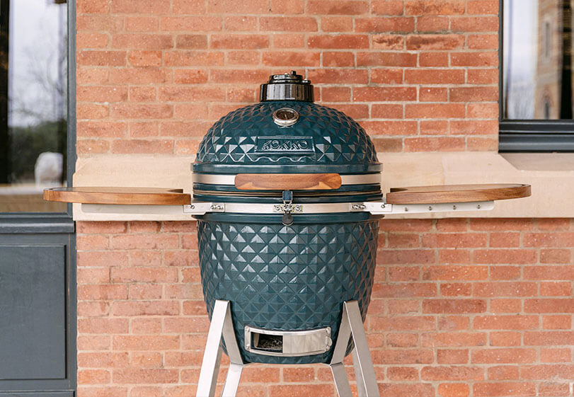 Kamado Kokko blu oceano posizionato su supporto con ripiani in legno, visto frontalmente all’esterno contro un muro di mattoni.