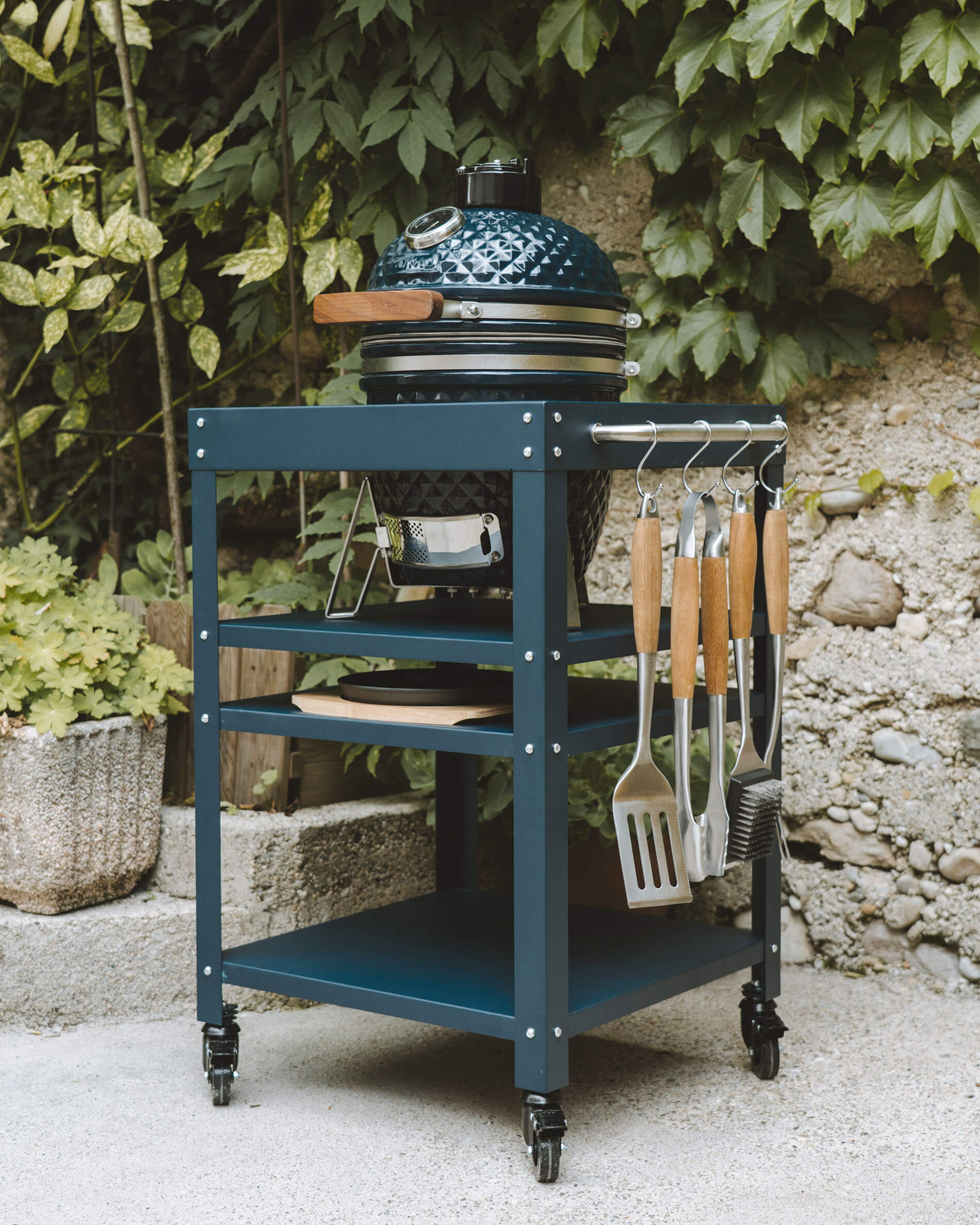 Kamado Kokko color blu oceano posizionato su un carrello metallico coordinato, con ripiani portaoggetti, cassetto raccogli-cenere e utensili appesi lateralmente.