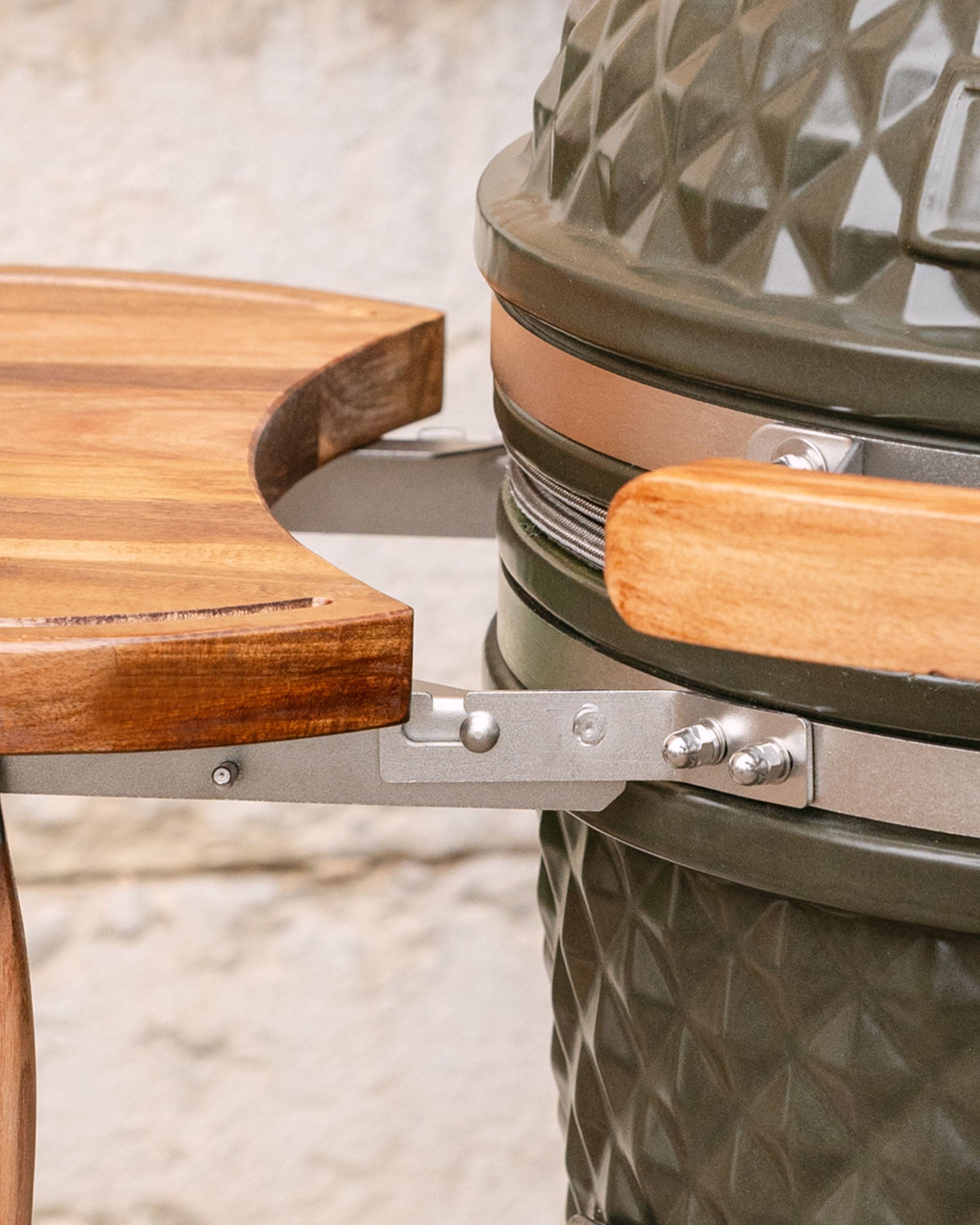Dettaglio dei fissaggi metallici che collegano il ripiano laterale in legno al kamado, con maniglia in legno e fascia in acciaio inox visibili.