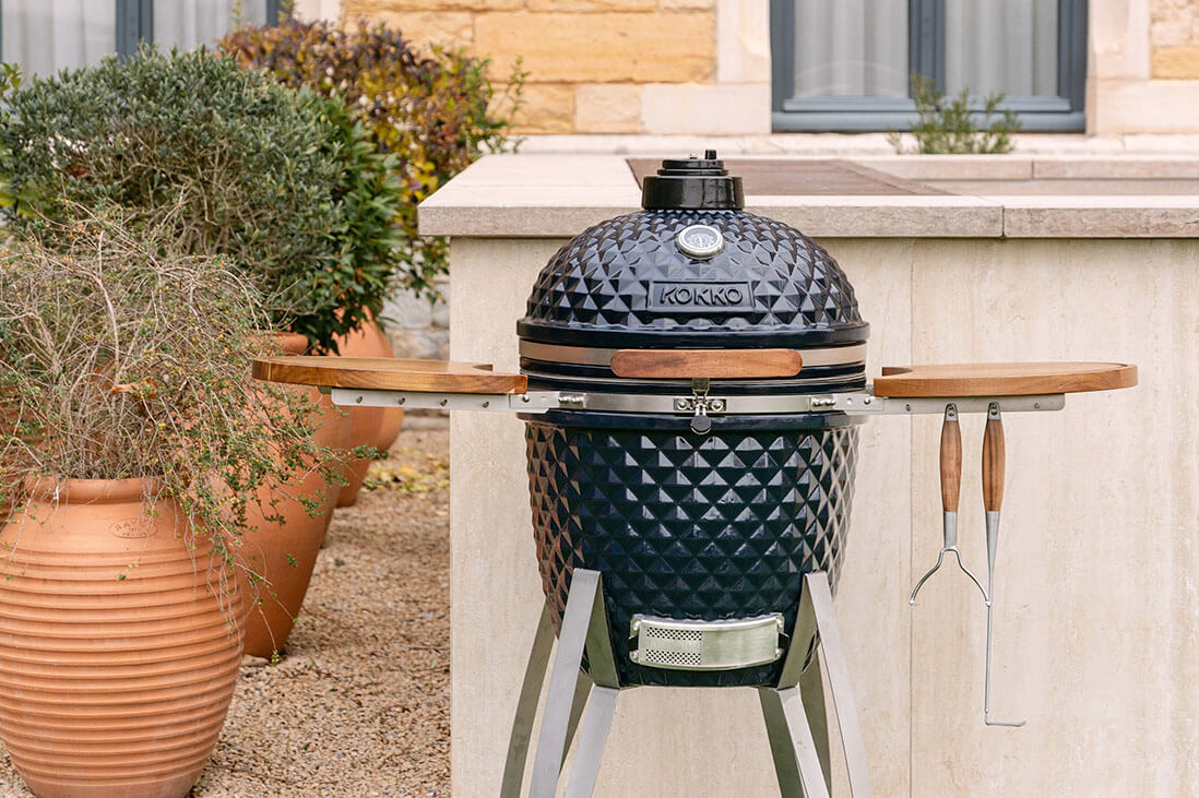Kamado Kokko posizionato all’esterno, con ceramica nera testurizzata, ripiani laterali in legno, utensili appesi e struttura in metallo, integrato in una cucina da giardino.
