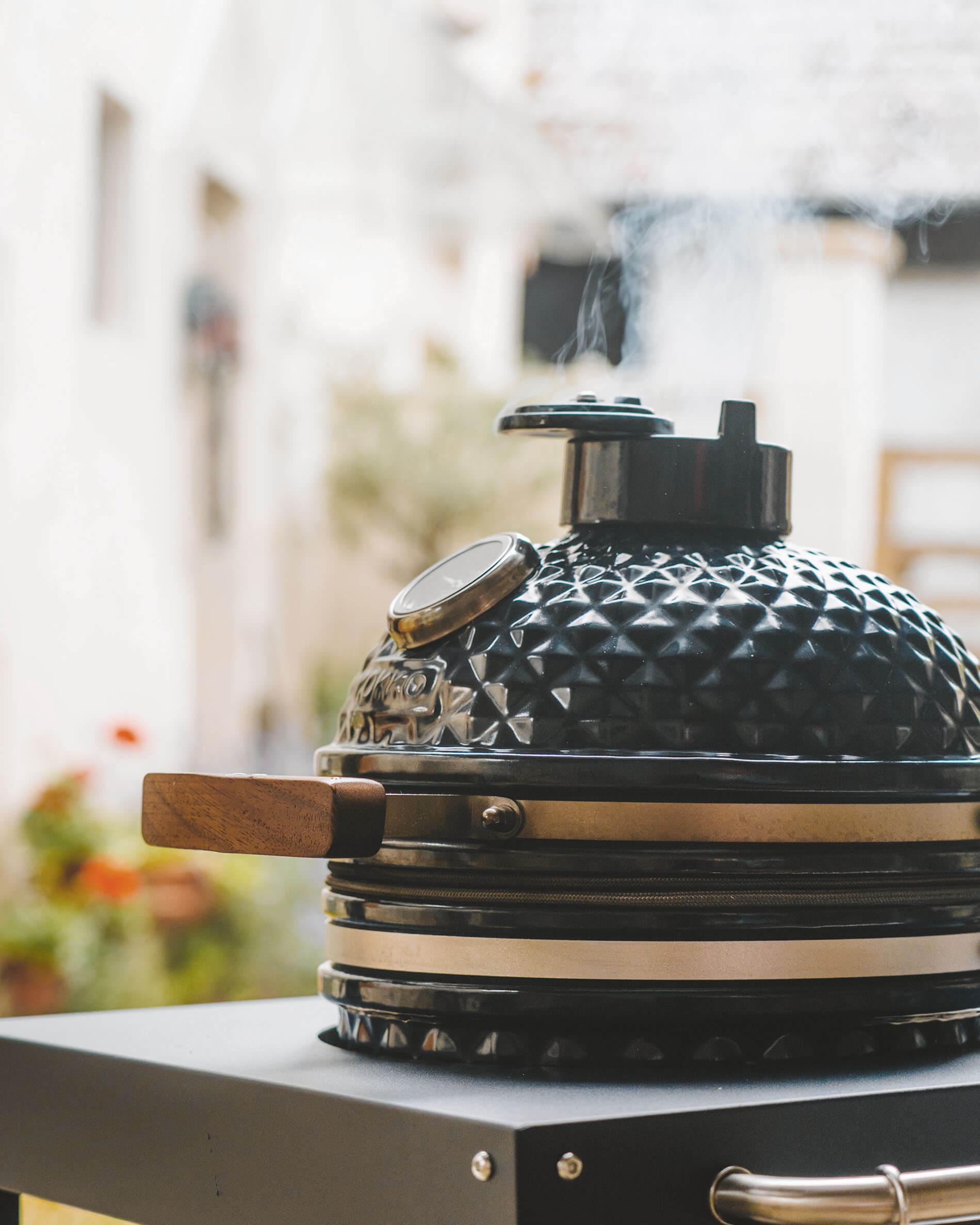 Kamado Kokko nero su carrello, con coperchio in ceramica testurizzata, maniglia in legno, termometro integrato e fumo che esce dal camino.