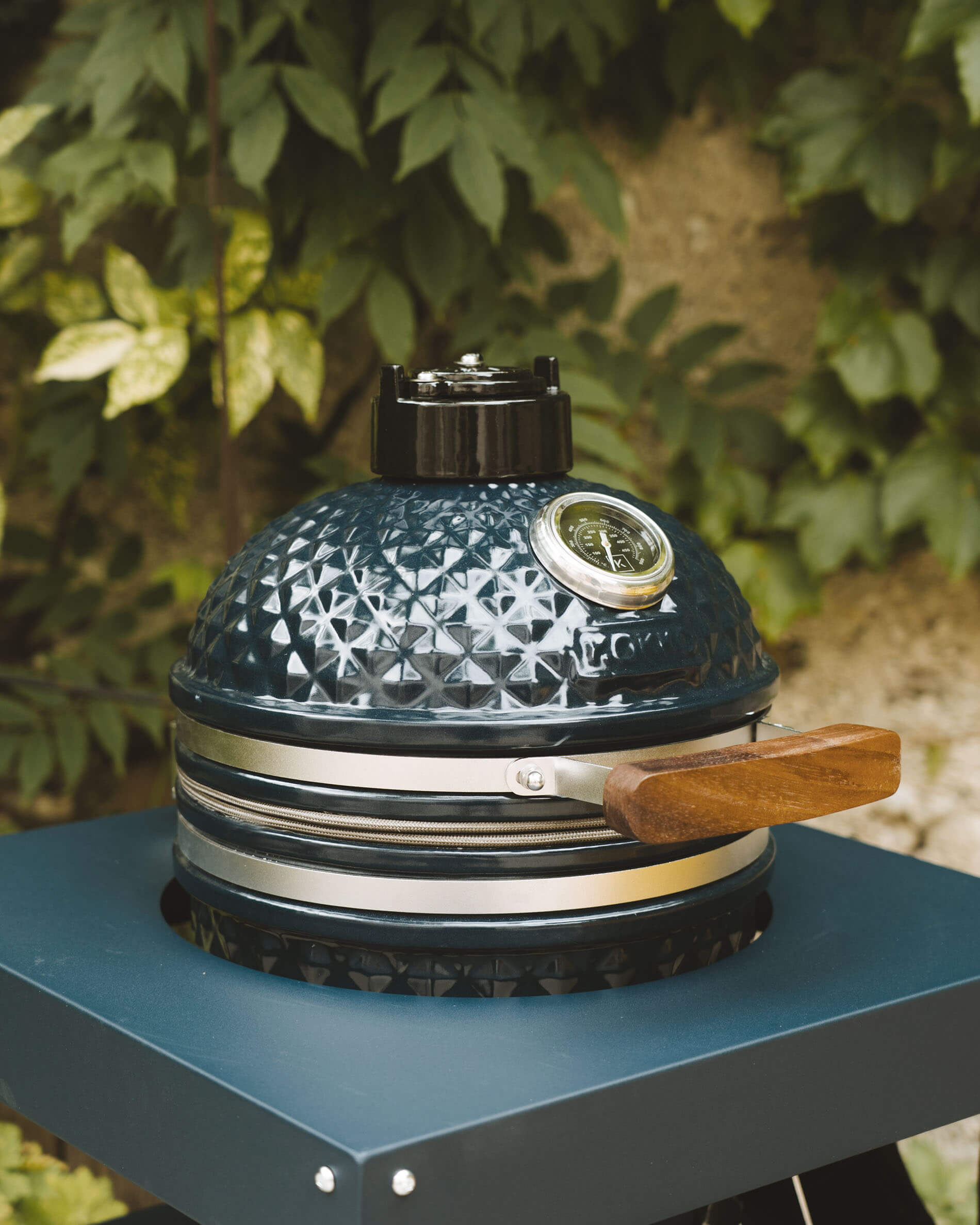 Vista frontale di un kamado Kokko color oceano su carrello, con ceramica testurizzata, termometro integrato e maniglia in legno in evidenza.