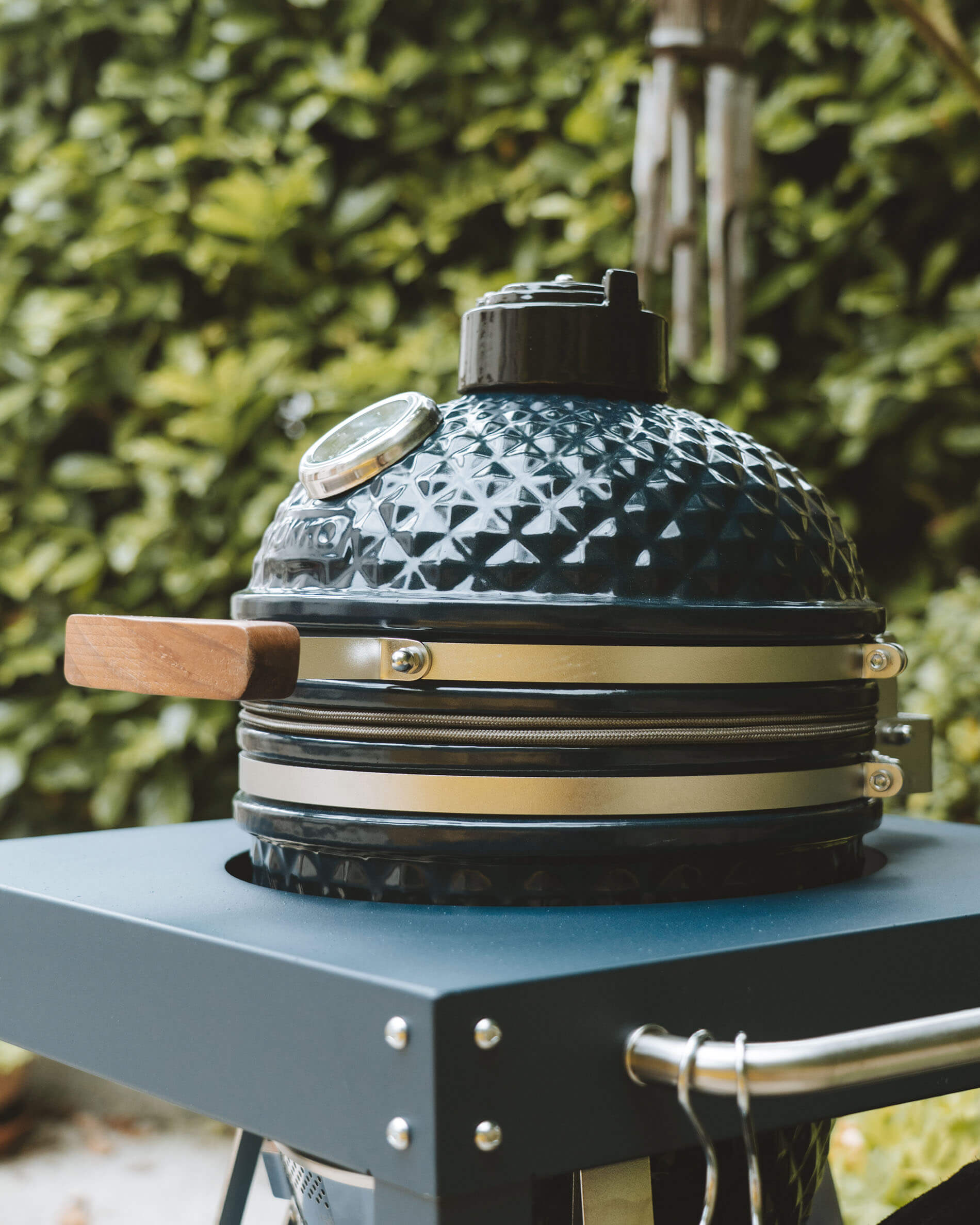 Vista laterale di un kamado Kokko color oceano su carrello, con coperchio in ceramica, maniglia in legno e termometro integrato in evidenza.