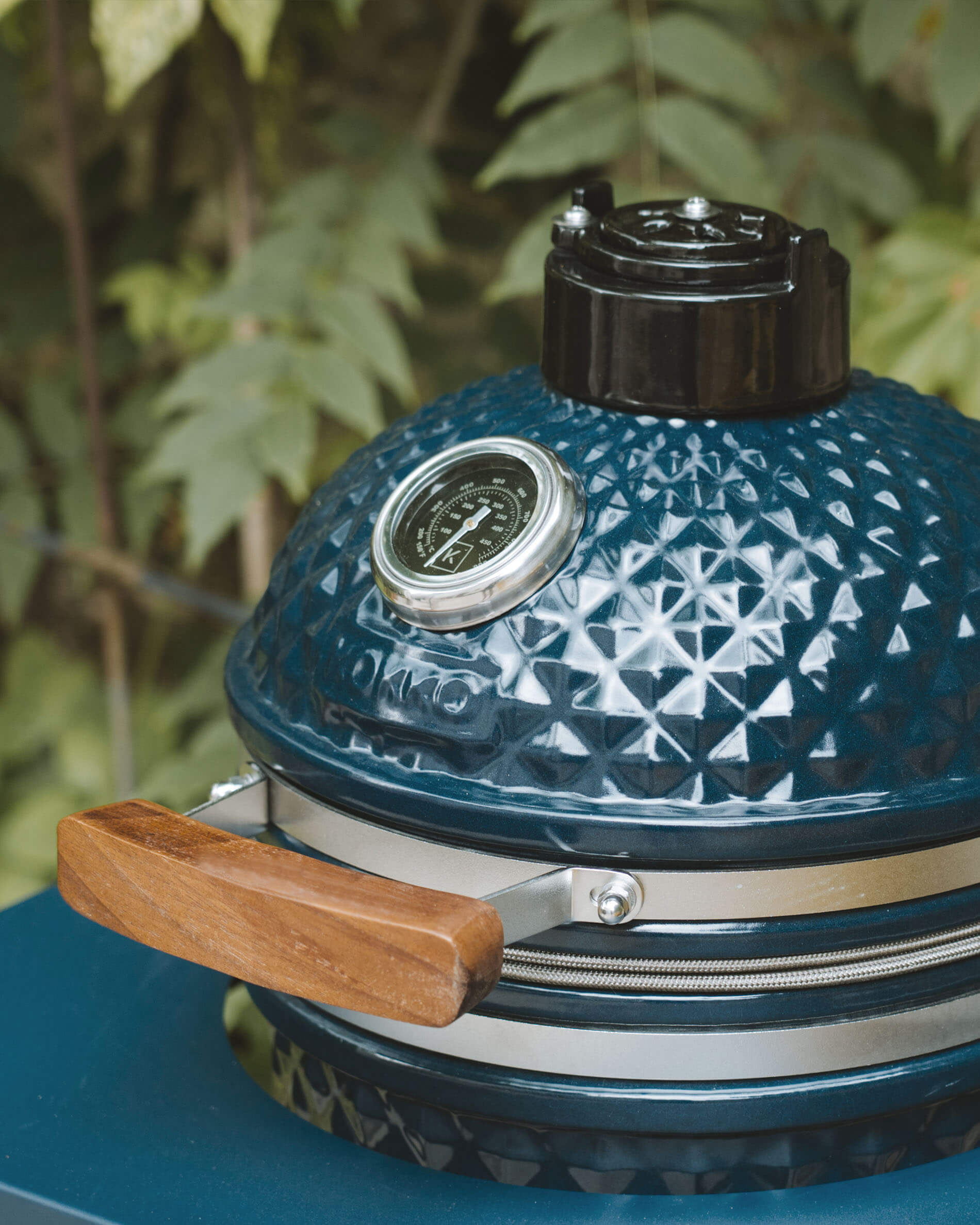 Dettaglio del coperchio di un kamado Kokko color oceano con termometro integrato e maniglia in legno.