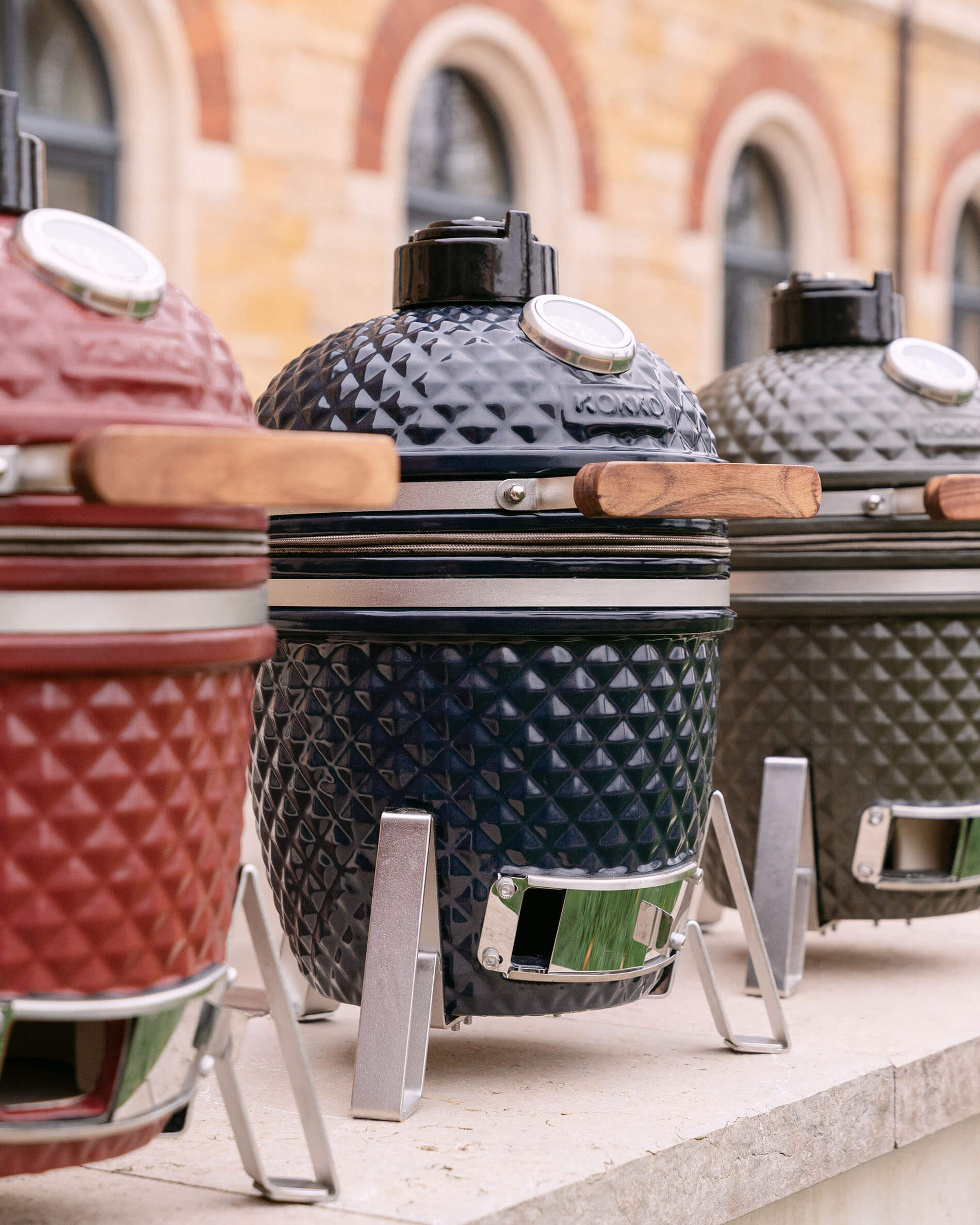 Kamado Kokko in ceramica testurizzata allineati, disponibili in diversi colori con manici in legno e supporti metallici.