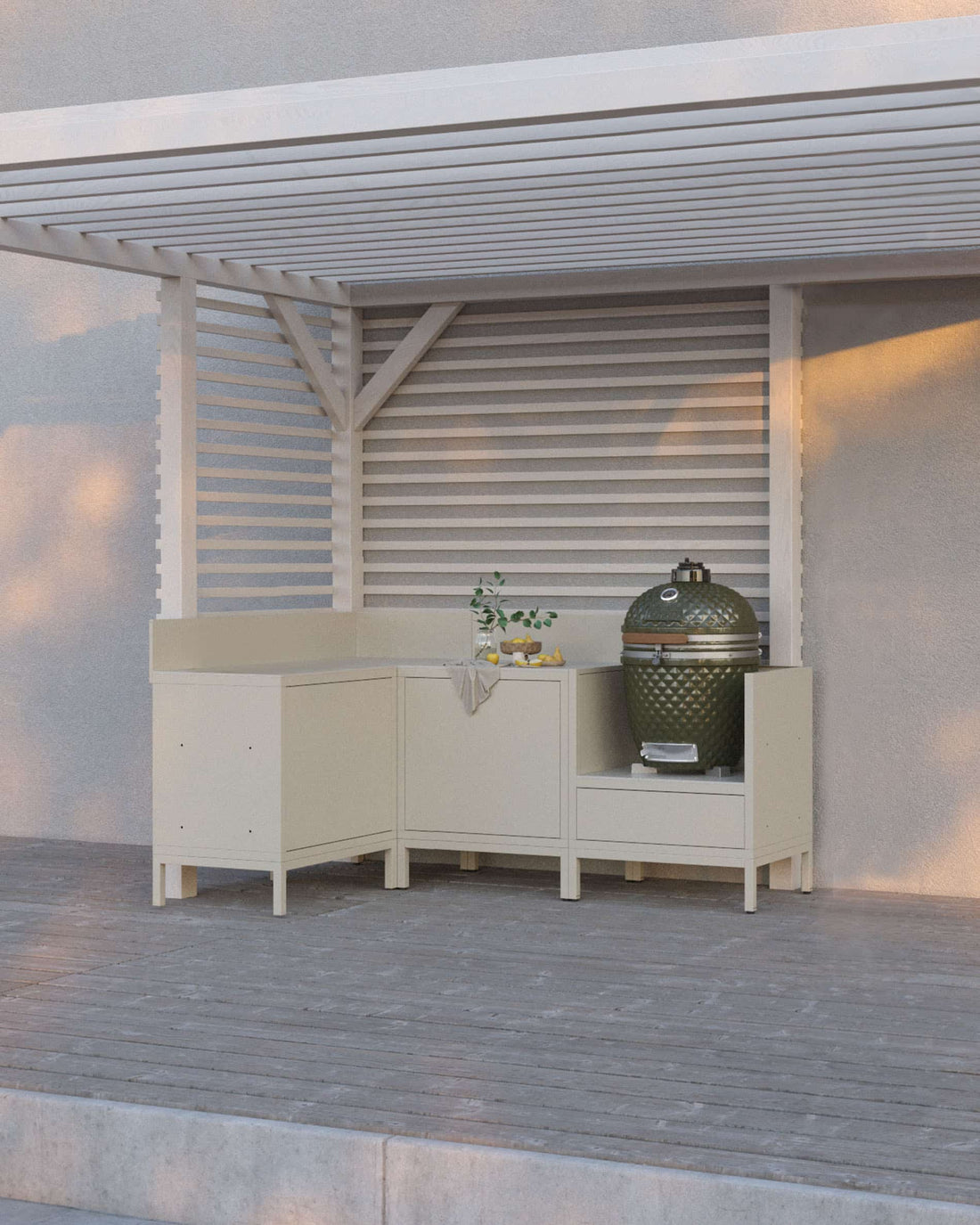 Cuisine pilotis coloris crème composée de quatre modules avec angle, plan de travail continu et emplacement dédié au kamado, installée sous une pergola blanche sur terrasse en bois.
