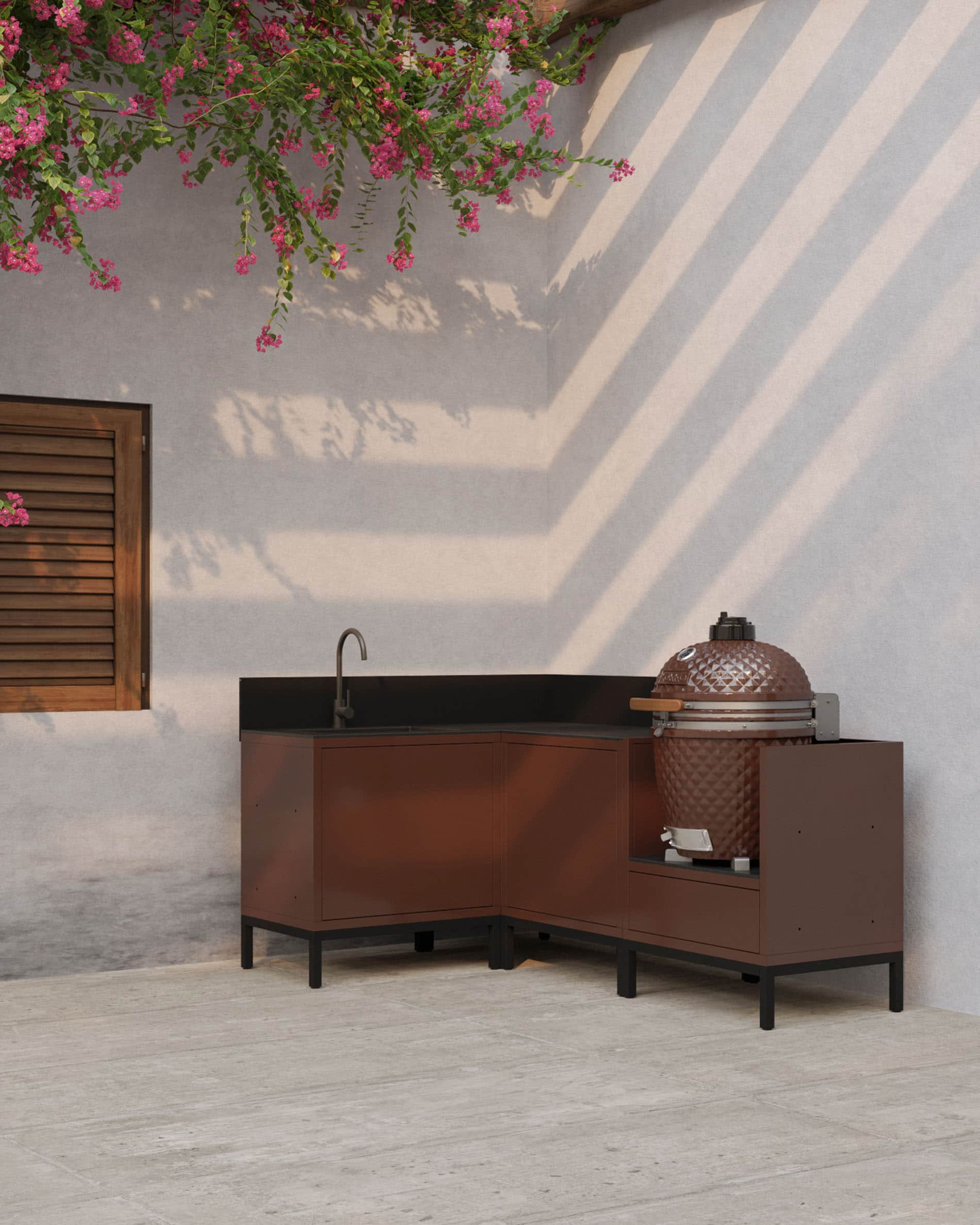Cucina su palafitte angolare color ruggine con piano di lavoro scuro, vani chiusi e modulo dedicato all’integrazione di un kamado per esterni.
